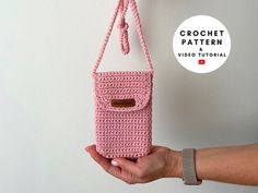 a hand holding a pink crocheted cell phone case hanging from a hook on a wall