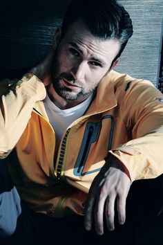a man sitting down with his hand on his head and looking at the camera while wearing a yellow jacket