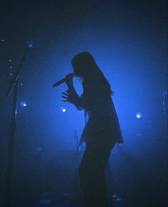 a woman standing in front of a microphone on stage with blue lights behind her and one arm raised up