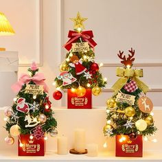 three small christmas trees in red boxes with gold and silver ornaments on them, decorated with lights