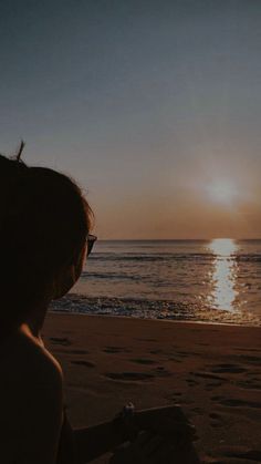 a woman sitting on top of a sandy beach next to the ocean at sun set