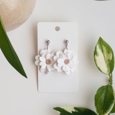 two white flower earrings sitting on top of a table next to green leaves and a potted plant