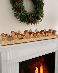 a christmas mantle with lit candles and houses on it next to a wreath that is hanging above the fireplace