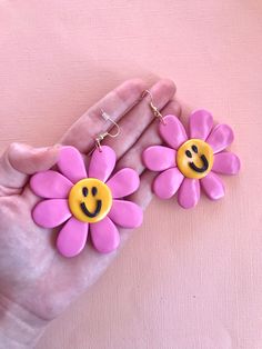 a pair of pink and yellow flower earrings with smiley face on the middle, in front of a pink background