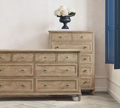 a wooden dresser with drawers and a flower vase on top in a white walled room