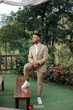 a man sitting on top of a wooden bench in a suit and white shoes with trees in the background