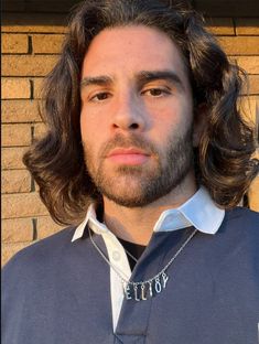 a man with long hair wearing a blue shirt and silver chain around his neck is standing in front of a brick wall