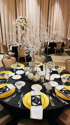 a black table topped with yellow plates and silverware next to a tall vase filled with white flowers