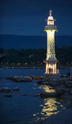 the lighthouse is lit up at night with people standing on it's rocks near the water