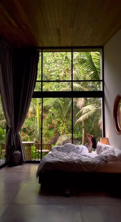 a person laying on a bed in front of a large window looking out onto the jungle