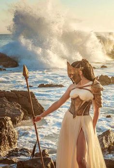 a woman in a white dress standing next to the ocean with a spear and shield