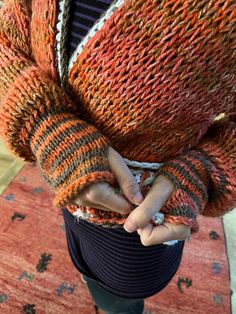 a woman is holding her hands in her pocket while standing on a rug with an orange and gray sweater