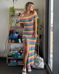 a woman in a multicolored crochet dress standing next to a window