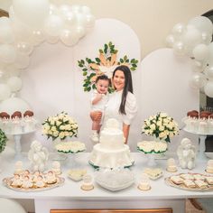 a woman holding a baby standing next to a table filled with cakes and desserts