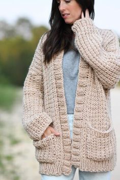 a woman wearing a cardigan sweater and jeans standing in front of the ocean with her hands on her hips