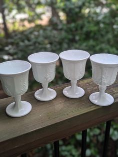 four white glass goblets sitting on top of a wooden table next to trees