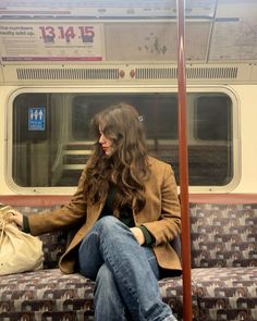 a woman with long hair sitting on a subway train holding a bag and looking at her phone