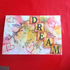 the word dream written in blocks with a bird on it and hearts around them, sitting on a red surface