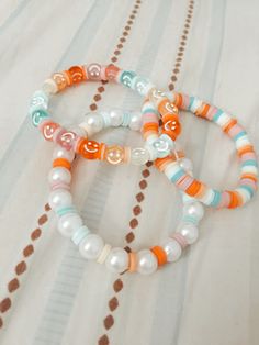 three bracelets with beads and charms on a white sheeted bed spread, one has an orange smiley face in the middle