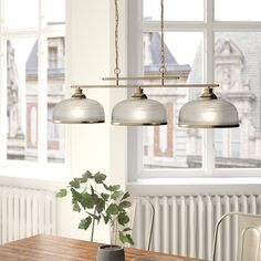 three lights hanging over a wooden table in front of a window with potted plant