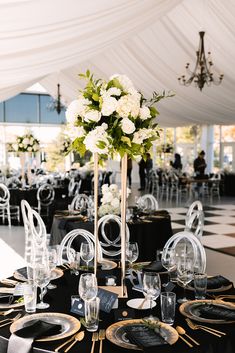 a table set up for an event with black linens and white flowers in a tall vase