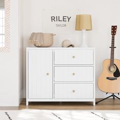 a white dresser with a guitar on top of it next to a lamp and rug