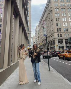 two women standing on the sidewalk talking to each other in front of some tall buildings