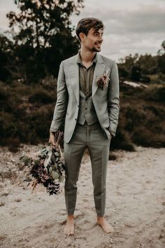 a man in a gray suit holding a bouquet and looking at the camera while standing on a beach