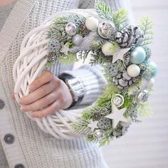 a woman is holding a wreath with ornaments on it and wearing a gray cardigan