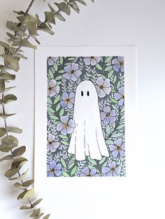 a card with a white ghost surrounded by leaves and flowers on a white wall next to a plant