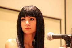 a woman with long black hair standing in front of a microphone and looking at the camera