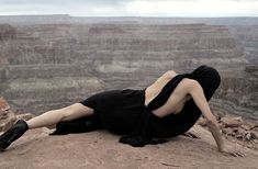 a woman laying on top of a cliff next to the ocean in a black dress