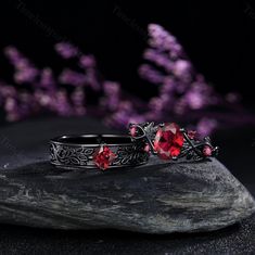 two rings with red stones sitting on top of a rock next to some purple flowers