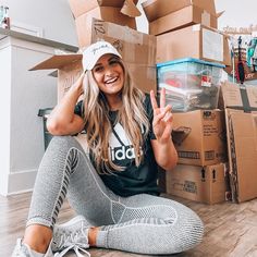 a woman sitting on the floor in front of boxes and giving the peace sign with her hand