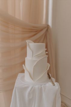 a white wedding cake sitting on top of a table next to a sheer drapes