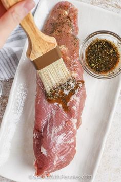 a person holding a brush over raw meat on a white platter with seasonings