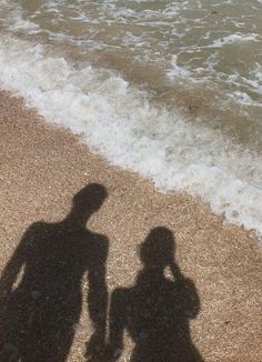 two people standing on the beach next to the ocean with their shadow in the sand