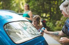 a woman is painting the side of a blue car with white lettering on it and two other women are behind her