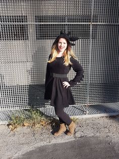 a woman standing in front of a fence wearing a black dress and hat with her hands on her hips