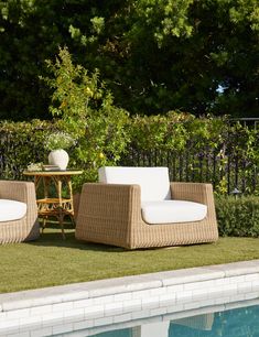 two wicker chairs sitting next to a small table near a swimming pool with green grass