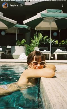 a woman wearing sunglasses sitting in a pool with an umbrellas over her head and looking at the camera