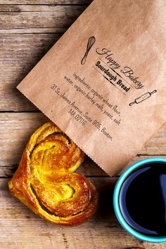 a pastry next to a cup of coffee on top of a wooden table with a brown paper bag