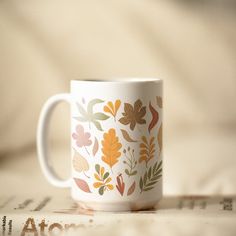 a coffee cup with leaves painted on it sitting on top of a table next to an open book