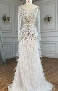 The White Rhinestone Beaded Sheer Long Sleeve Feather Embellished Backless Gown is a stunning and ethereal piece, perfect for making a grand entrance at any formal event. This gown features sheer long sleeves adorned with delicate rhinestone beading, adding a touch of sparkle and elegance. The bodice and back are also embellished with rhinestones, creating a dazzling effect. The gown's backless design adds a bold and dramatic element, while feather embellishments along the skirt provide a luxuri White Embellished Evening Dress For Reception, Glamorous White Wedding Dress For Reception, Elegant Evening Wedding Dress With Rhinestones, Glamorous White Embellished Evening Dress, White Glamorous Embellished Evening Dress, Luxury Embellished Floor-length Wedding Dress, Fitted Evening Dress With Rhinestone Fringe For Wedding, Elegant Rhinestone Fringe Evening Dress For Prom, Glamorous White Evening Dress With Rhinestones