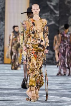 a woman walking down a runway wearing a dress with flowers on it and holding a cane