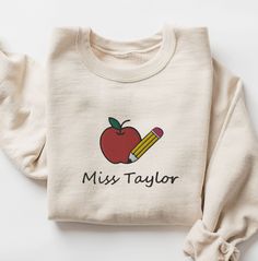 These soft unisex Comfort Colors shirts are embroidered with an apple and pencil along with your custom teacher's name :)  * 100% ring-spun cotton * Fabric weight: 6.1 oz/yd² (206.8 g/m²) * Garment-dyed * Relaxed fit * 7/8″ double-needle topstitched collar * Twill-taped neck and shoulders for extra durability * Double-needle armhole, sleeve, and bottom hems SIZING:  They're unisex but fit true to size (or very close to) for ladies. We recommend sizing up though if you prefer a roomier or oversized look! Guys, we definitely suggest sizing up. As these shirts are personalized we do not offer exchanges or refunds unless an error has been made so please check measurement chart before ordering. We would love to assist if you need help! CARE: Wash inside out in cold water and hang to dry or dry Personalized Cotton T-shirt For Fall, White Long Sleeve Tops For Back To School, Customizable Crew Neck Sweatshirt For School, School Long Sleeve T-shirt With Name Print, School Spirit Cotton Top With Custom Embroidery, Cotton Sweatshirt With Name Print, Cotton Tops With Custom Embroidery For School Spirit, Personalized Crew Neck Top For College, School T-shirt With Embroidered Crew Neck