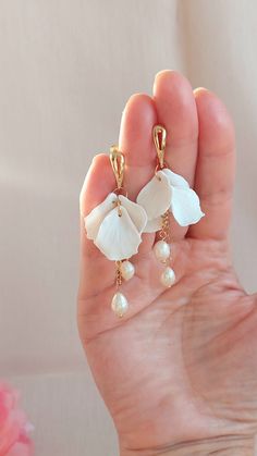 a person's hand holding two white flower and pearl drop earrings in front of a pink flower