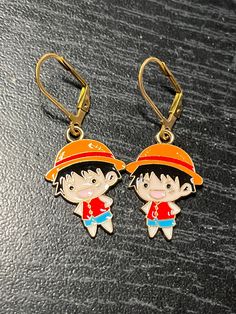 two cartoon characters are attached to gold colored earwires on a black surface, one is wearing an orange hat and the other has a red shirt