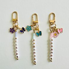 three key chains with flowers and name charms attached to them, on a white surface