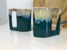 two coffee mugs sitting next to each other on a white counter top in front of a wicker basket